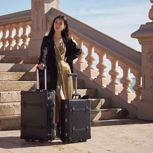 A woman stands on stone stairs beside the sea, holding two hard-shell black rolling suitcases. She wears a black blazer over a gold dress.