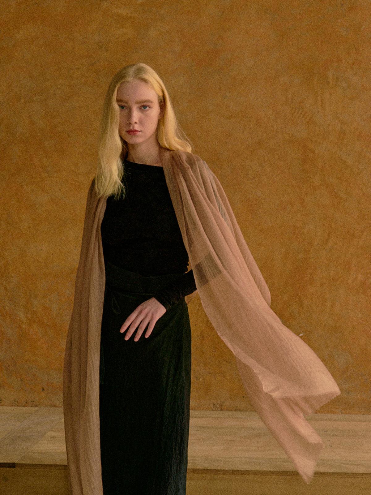 A blonde woman stands against a textured warm tan wall wearing a black velvet long-sleeve top and black pleated skirt, with a sheer blush-beige draped wrap or cape flowing around her.