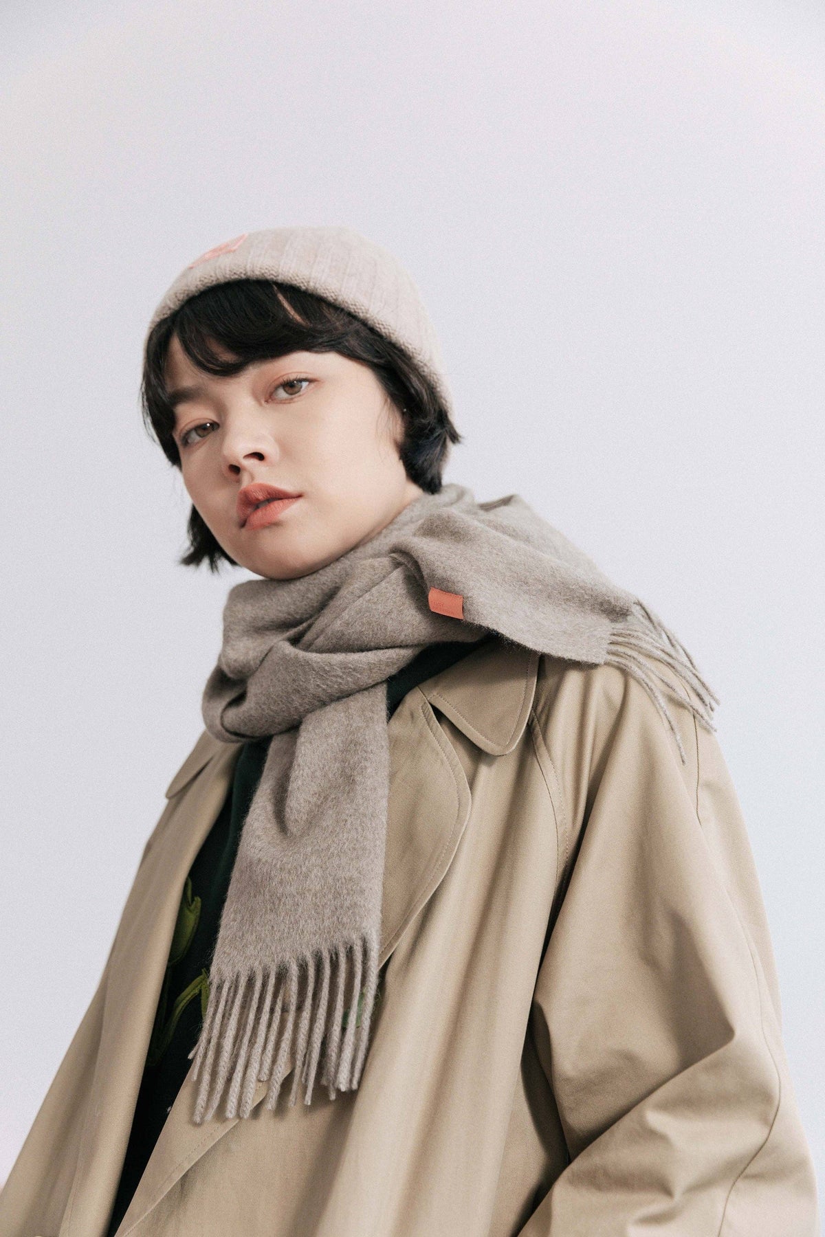 A model wearing a beige knit beanie, a thick gray wool scarf with fringe, and a beige trench coat, photographed against a light gray studio background.