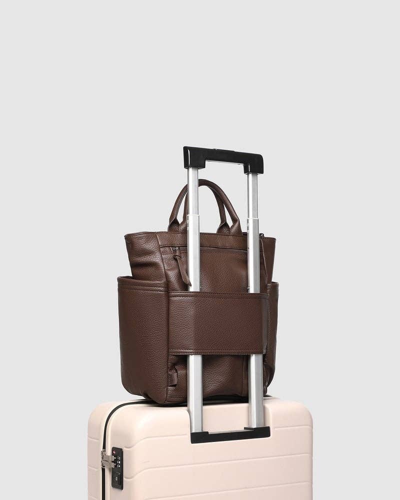 A brown pebbled leather tote bag with silver telescoping handles, shown resting on top of a light pink hard-shell suitcase against a light gray studio background.