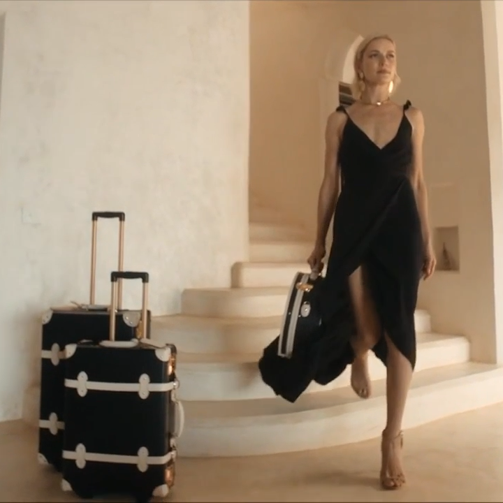 A woman in a black sleeveless dress walks down a staircase while carrying a black duffel bag, with a black hard-shell suitcase standing beside her in a bright, modern interior.