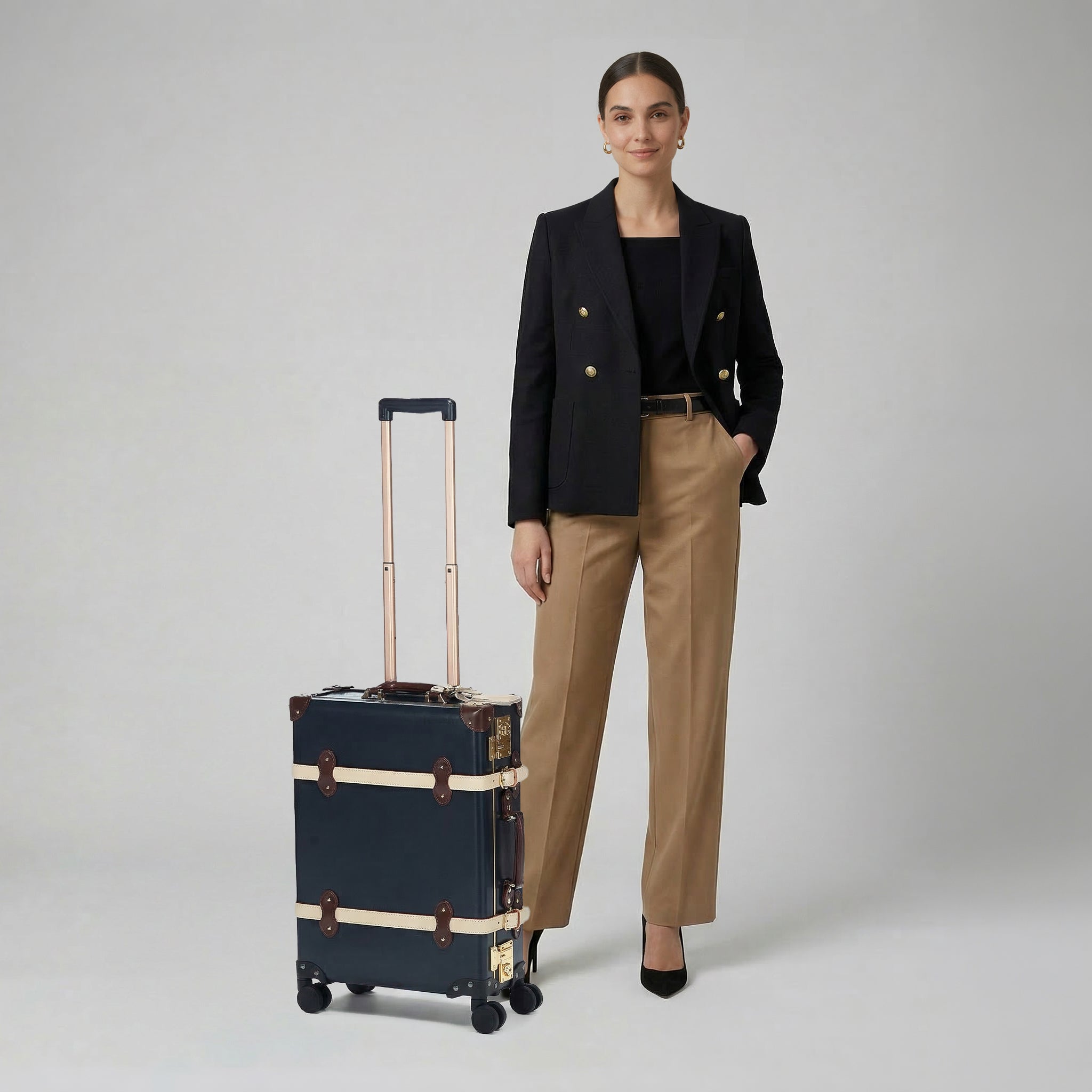 Woman in black blazer and beige pants standing next to a dark suitcase on a light gray background
