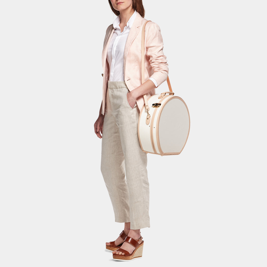 Woman holding a round white bag wearing a light pink blazer and beige pants on a white background