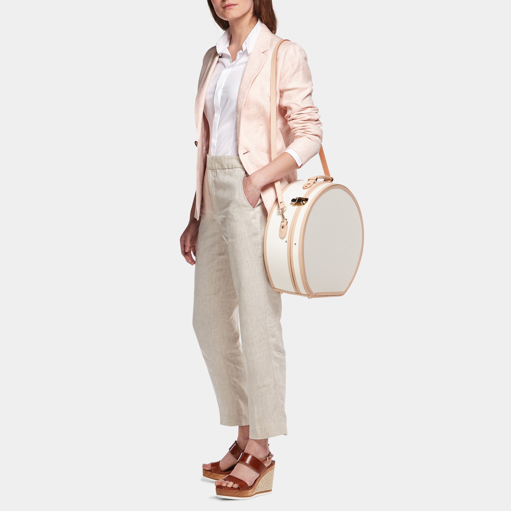 Woman holding a round white bag wearing a light pink blazer and beige pants on a white background