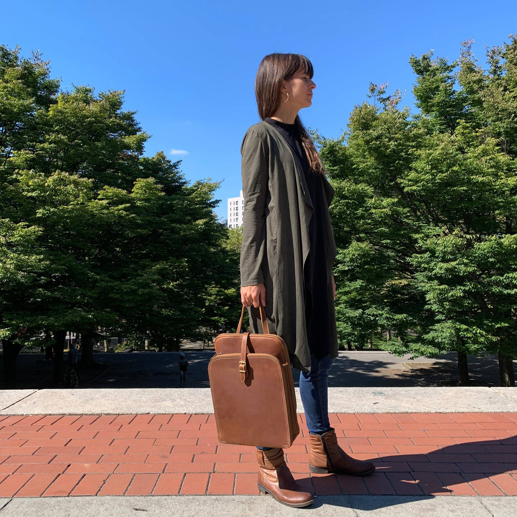 Person holding a brown leather briefcase outdoors with trees and blue sky in the background