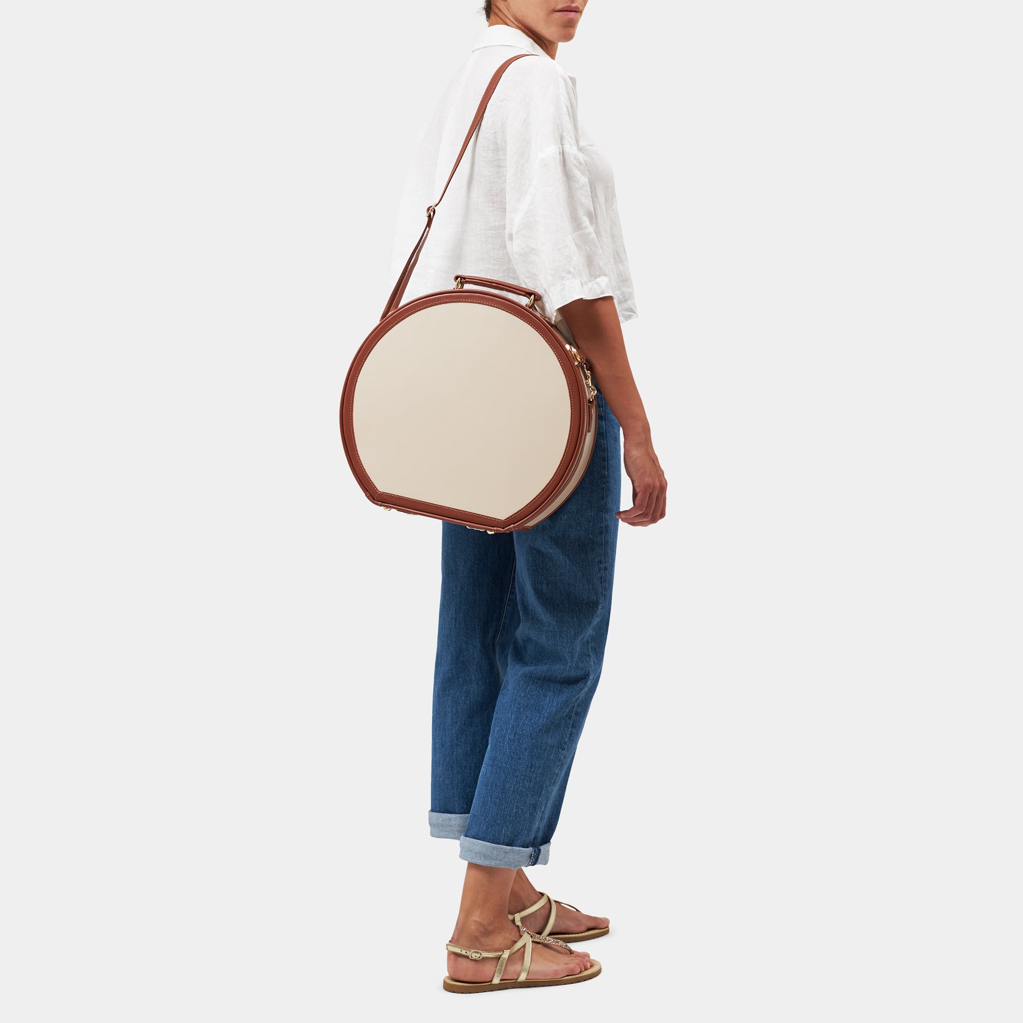 Person holding a round cream leather hatbox with brown leather trim on a white background