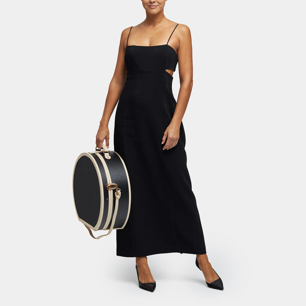 Woman in a black dress holding a black and beige round suitcase on a white background