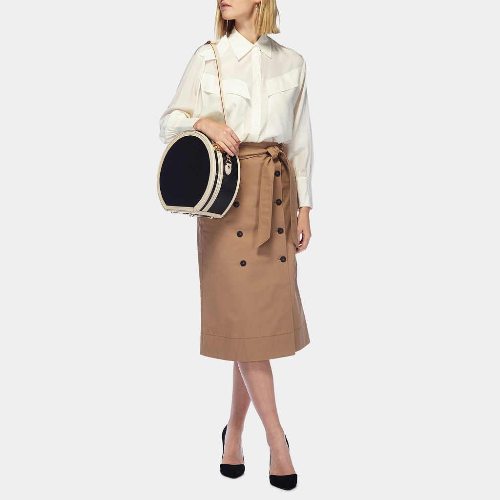 Woman holding a round black and beige bag wearing a white shirt and brown skirt with black buttons on a white background