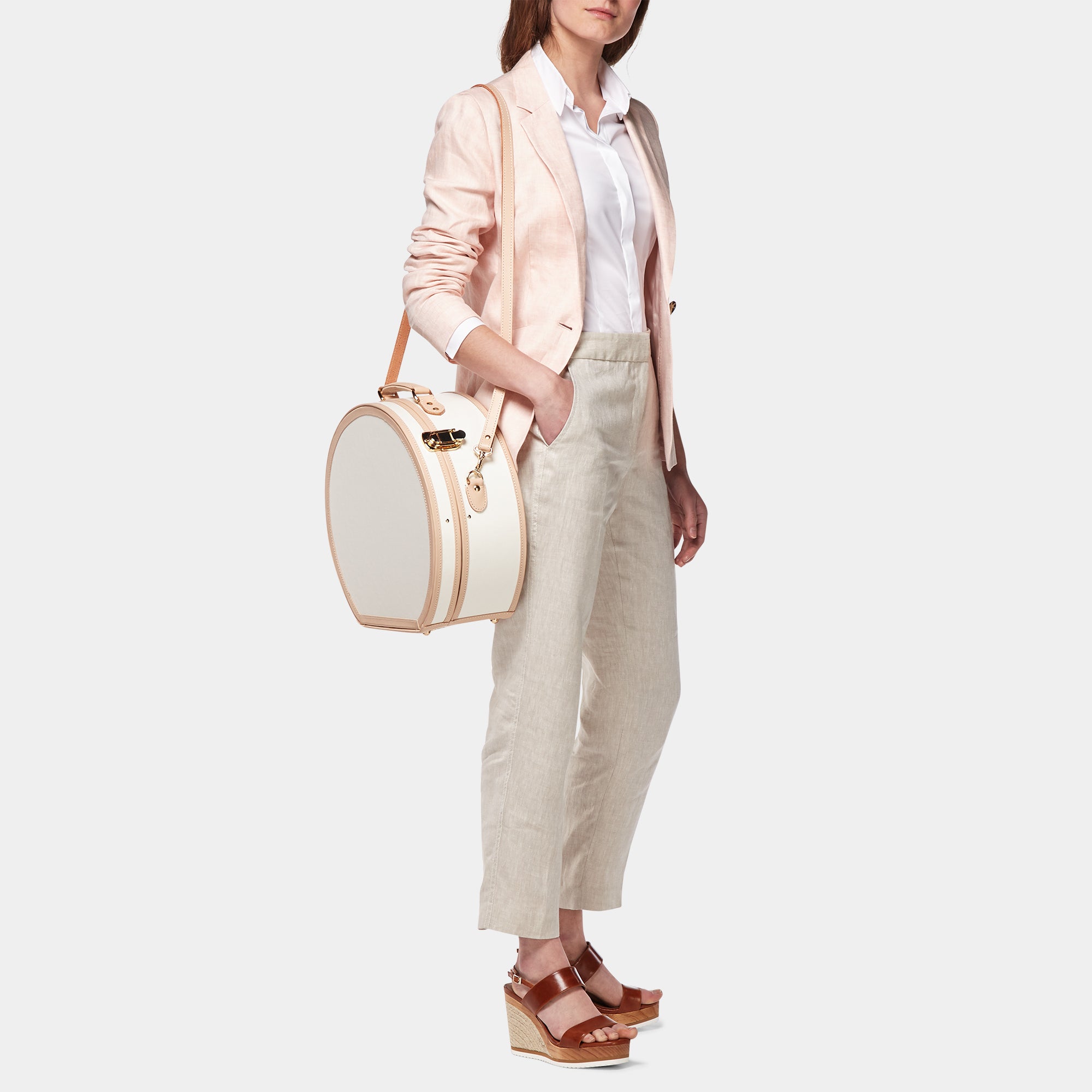 Woman holding a round beige handbag wearing a light pink blazer and white pants on a white background